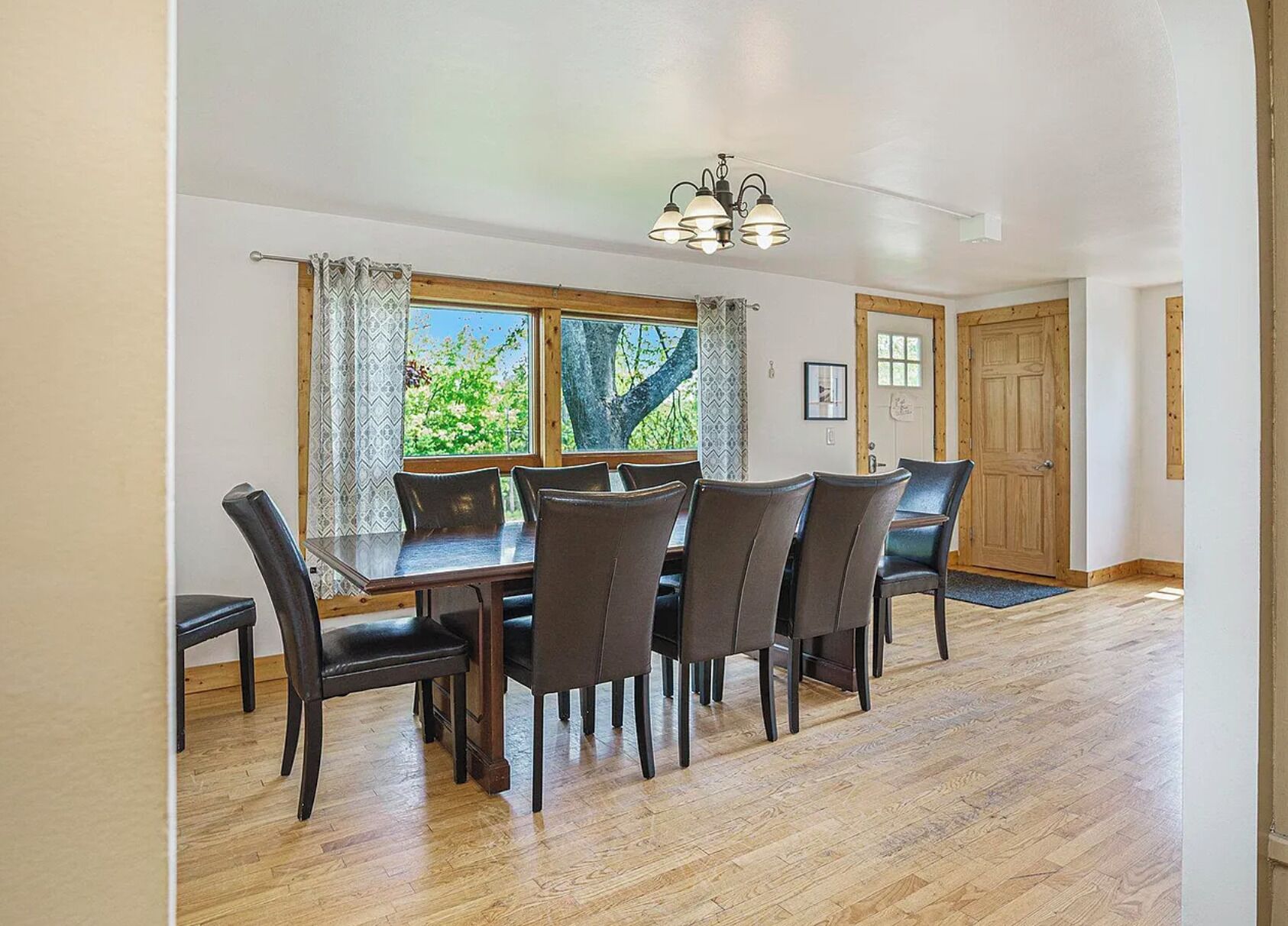 A dining area with a wooden table and chairs, a chandelier, and a view of greenery outside.