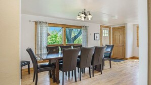 A dining area with a wooden table and chairs, a chandelier, and a view of greenery outside. - Lovely Lakehouse by Grand Traverse Bay. Pet Friendly! (Traverse City)