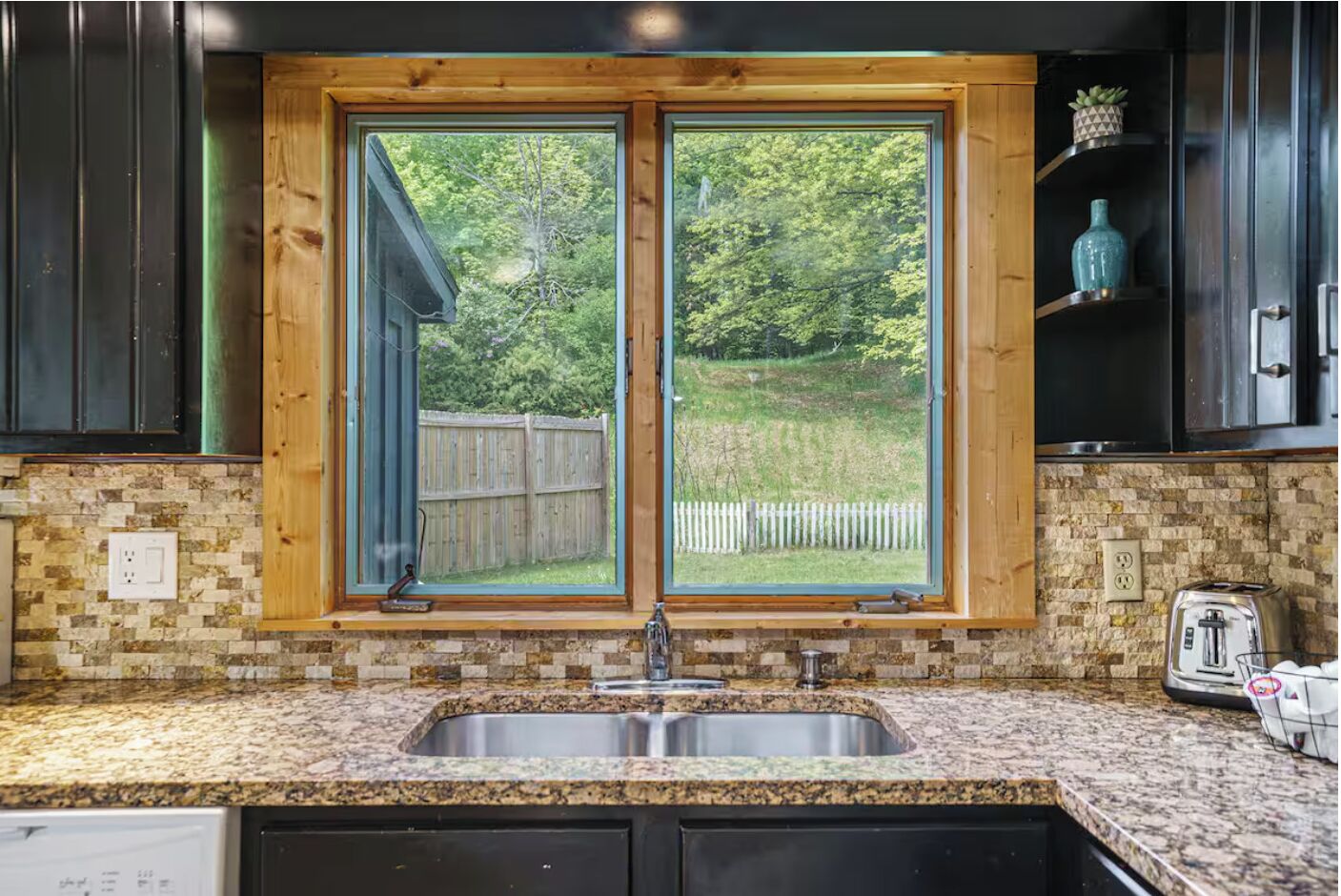 A kitchen with a granite countertop, a double sink, and a window with a view of a wooden deck and trees.