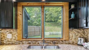 A kitchen with a granite countertop, a double sink, and a window with a view of a wooden deck and trees. - Lovely Lakehouse by Grand Traverse Bay. Pet Friendly! (Traverse City)