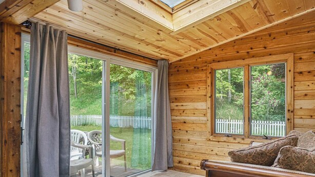 A wooden cabin interior with a sliding glass door leading to a deck, a sofa, and a view of a white picket fence and trees. - Lovely Lakehouse by Grand Traverse Bay. Pet Friendly! (Traverse City)