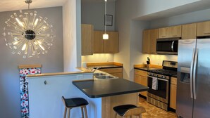 A modern kitchen with wooden cabinets, a stainless steel refrigerator, a built-in oven, and a unique ceiling light fixture. - SUNSET PLACE (Grand Haven)