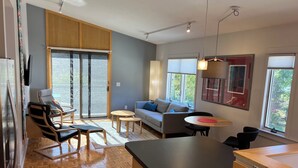 A modern kitchen with a large island, a dining area with a round table, and a sofa in a bright living space with large windows. - SUNSET PLACE (Grand Haven)
