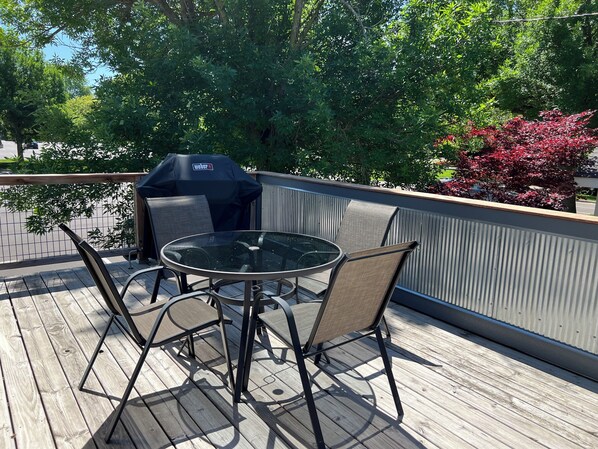 An outdoor deck with a round glass table and metal chairs, a grill, and a view of trees and a red-leaved plant. - SUNSET PLACE (Grand Haven)