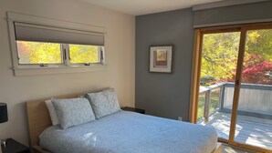 A bedroom with a bed, a nightstand, a window with blinds, a framed picture on the wall, and a view of a deck with autumn foliage. - SUNSET PLACE (Grand Haven)