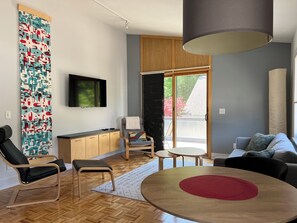 A modern living room with a flat-screen TV, a sofa, a dining table, and a view of the outdoors. - SUNSET PLACE (Grand Haven)