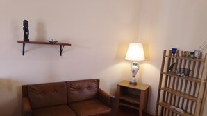Un salón con un sofá de cuero marrón, una mesita de madera con una lámpara, una estantería con objetos decorativos y un mueble tipo estante de madera a rayas.