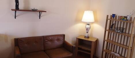 A living room with a brown leather sofa, a small wooden table with a lamp, a shelf with decorative items, and a striped wooden shelf unit.