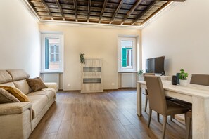 A living room with a sofa, a dining table, a TV, and two windows with shutters.