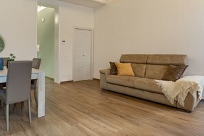 A room with a wooden floor, a beige sofa with a throw blanket, a dining table with chairs, and a white door.