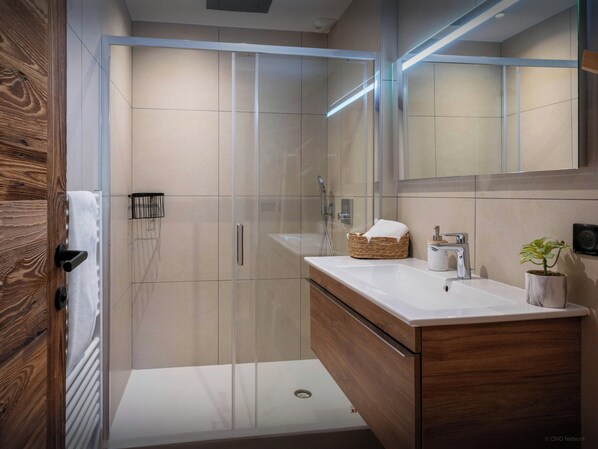Une salle de bain moderne avec une douche à l’italienne en verre, un meuble-lavabo en bois avec un plan de travail blanc et un grand miroir au-dessus du lavabo.