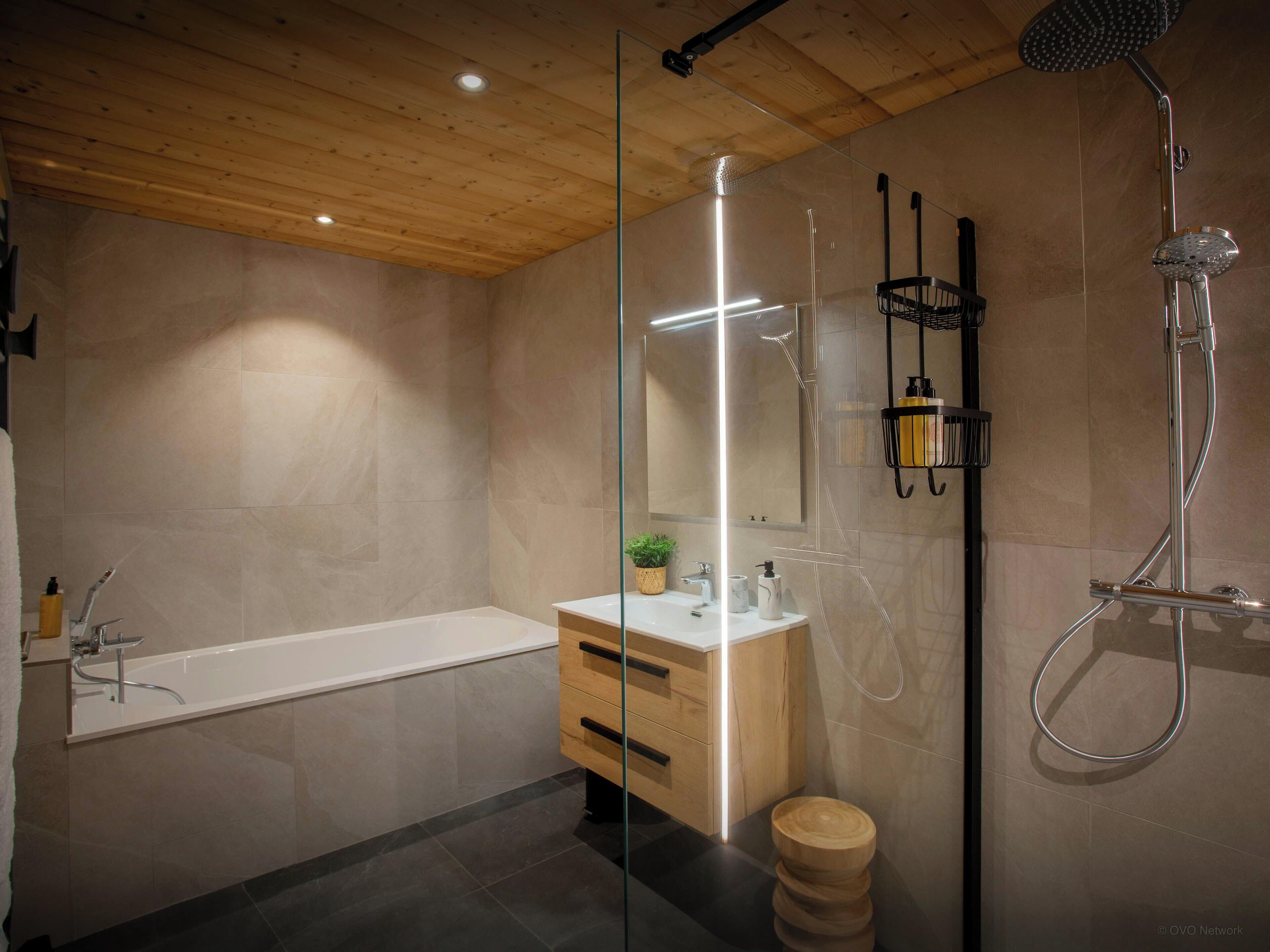 A modern bathroom with a wooden ceiling, a glass shower enclosure, a white bathtub, a wooden vanity, and a mirror.