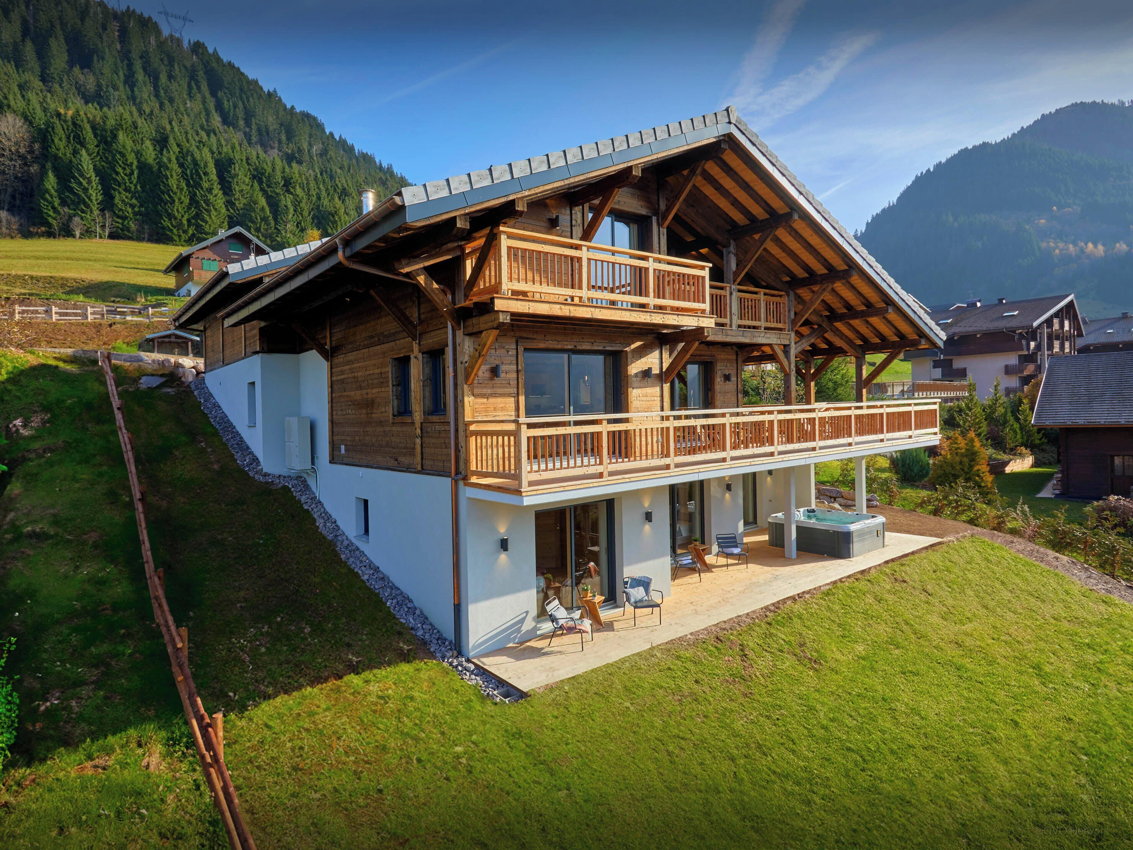 A two-story wooden house with a balcony, a patio area with seating, and a view of a mountainous landscape.