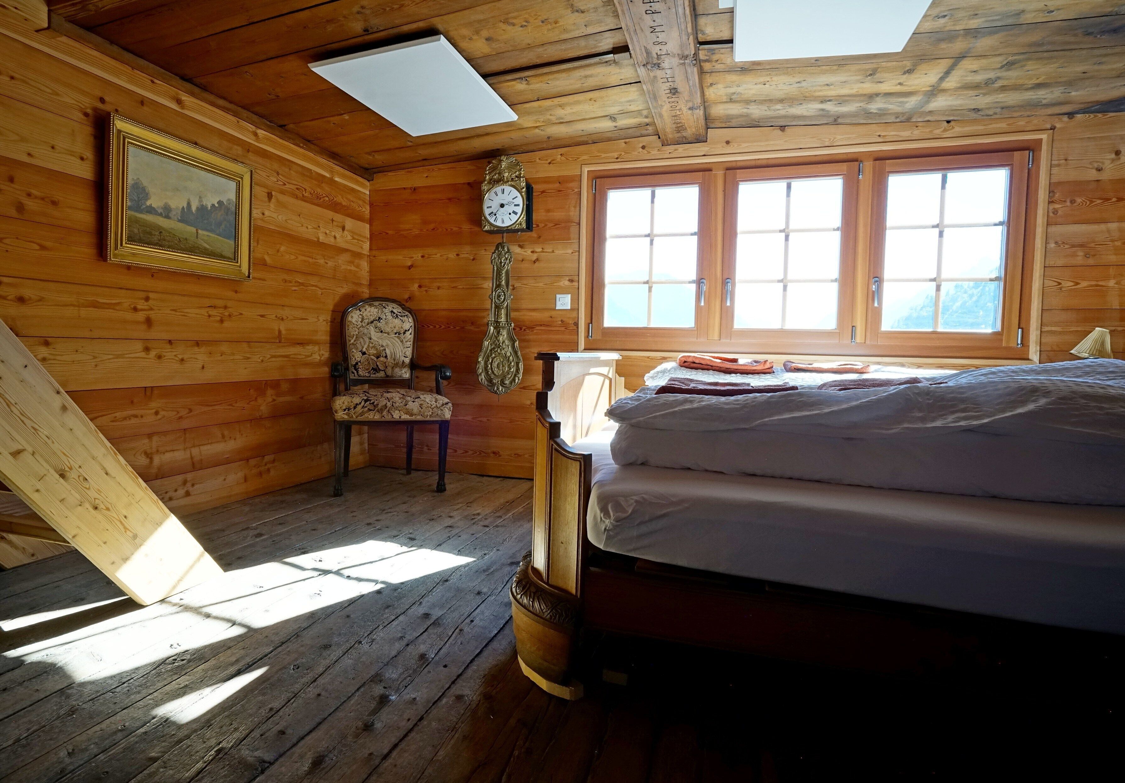 A wooden cabin room with a bed, a chair, a wall clock, and a window.