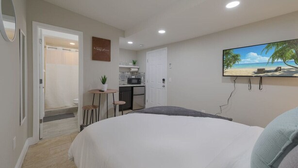 A bedroom with a large bed, a desk with a chair, and a small table with a plant. There is a bathroom with a shower and a toilet visible through an open door.