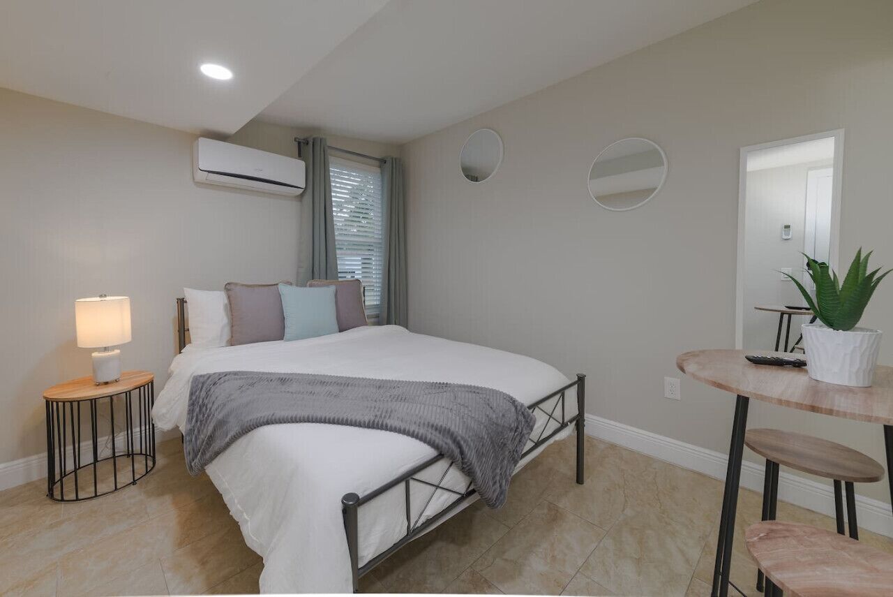 A bedroom with a bed, bedside lamp, air conditioning unit, window with blinds, and a small table with a potted plant.