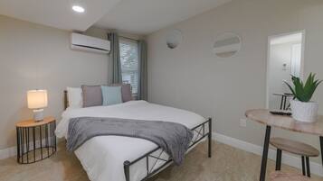 A bedroom with a bed, bedside lamp, air conditioning unit, window with blinds, and a small table with a potted plant.