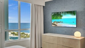 A hotel room with a large window offering a view of the ocean, a flat-screen TV mounted on a textured wall, and a bedside table with a lamp.