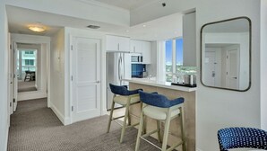 A modern kitchen with white cabinets, a stainless steel refrigerator, and a microwave. There are two bar stools with blue cushions. A large mirror is mounted on the wall.