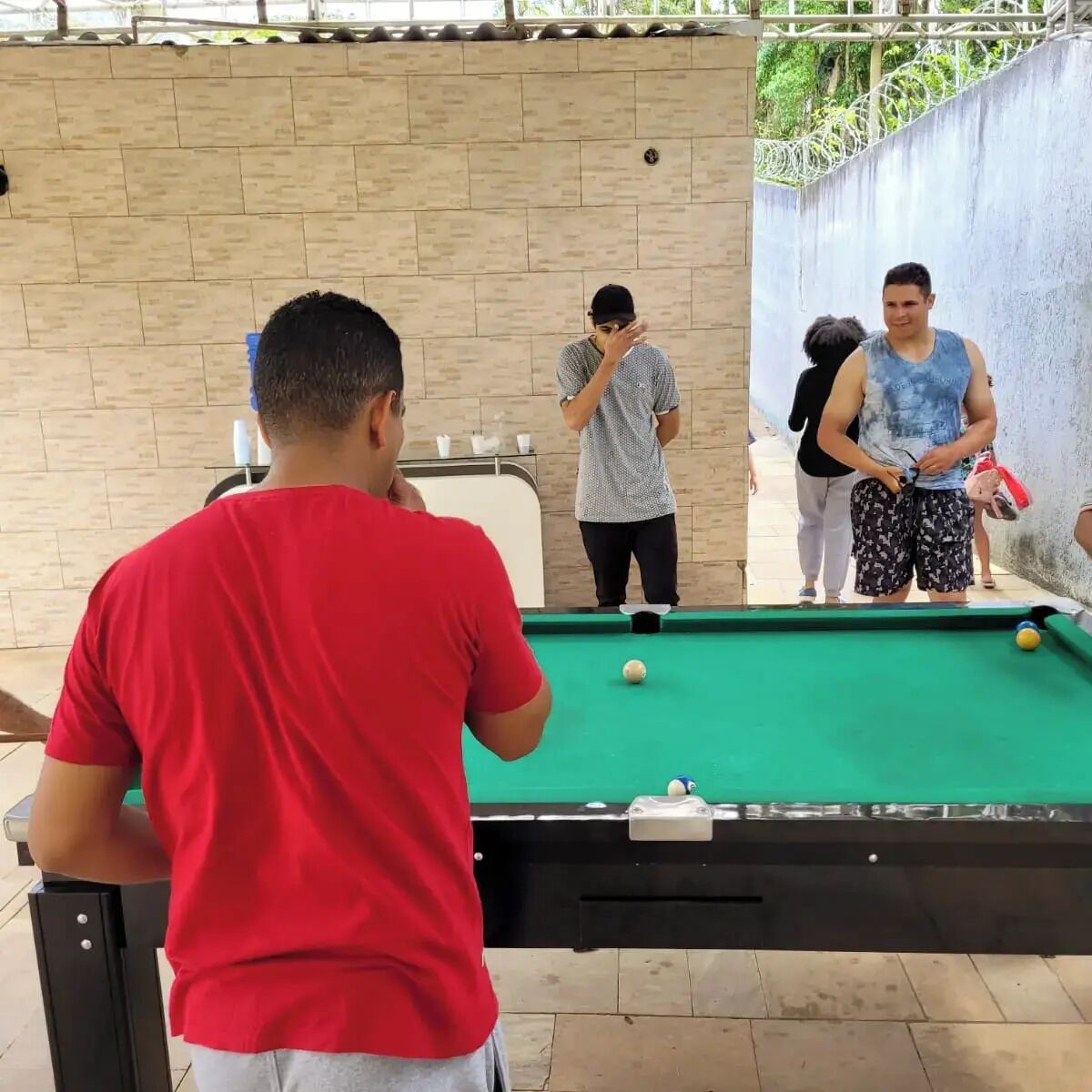 A pool table with a green felt surface, a white cue ball, and a yellow ball. There are two people standing at the table, one holding a cue stick. The background includes a brick wall and a metal railing.