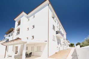 A multi-story white building with balconies, a covered outdoor seating area, and a clear sky.