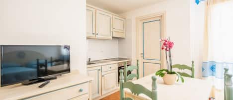 Cocina compacta con televisor, armarios de madera y zona de comedor con mesa y sillas.