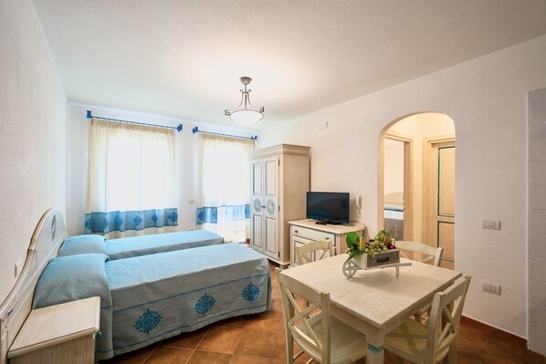 A room with two beds, a TV, a dining table, and a small kitchen area.