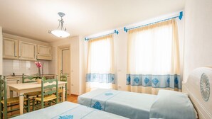 Una habitació amb dos llits, taula de dinar amb caires, zona de cuina i finestra amb cortines.