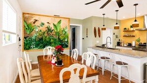 Un coin repas avec une table en bois et des chaises blanches, une cuisine dotée d’un îlot central et un ventilateur de plafond.
