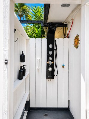 Un área pequeña y privada para ducha al aire libre, con puerta de madera blanca, manguera negra y panel de control de agua.