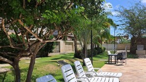 Un patio con sillones blancos y una mesa, rodeado de vegetación y un edificio al fondo.
