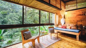 A wooden interior with a large window overlooking a forest, a bench, and a small table. - Deluxe Twin Room (Cahuita)