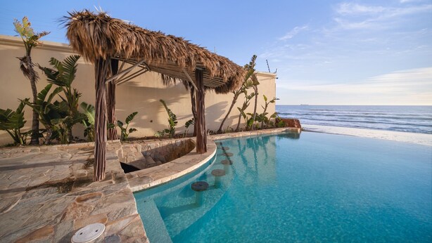 A poolside area with a thatched roof structure, stone steps leading into the water, and a view of the ocean. - Luxury Ocean Front Modern Condo in Rosarito BC (Rosarito)
