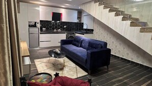 Una sala moderna con un sofá, una mesa de centro y una silla roja. Se observa una cocina al fondo con salpicadero blanco y negro.