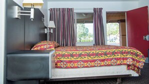 A hotel room with a bed, a bedside lamp, a window with curtains, and a red door. - Travelers Rest Motel (San Jose)