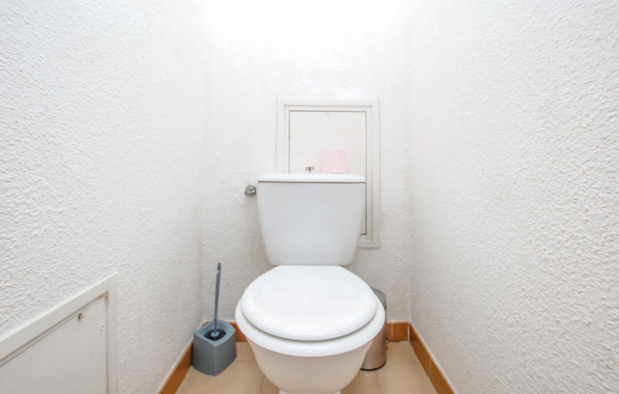 A small, white bathroom with a toilet, a mop, and a small trash bin.