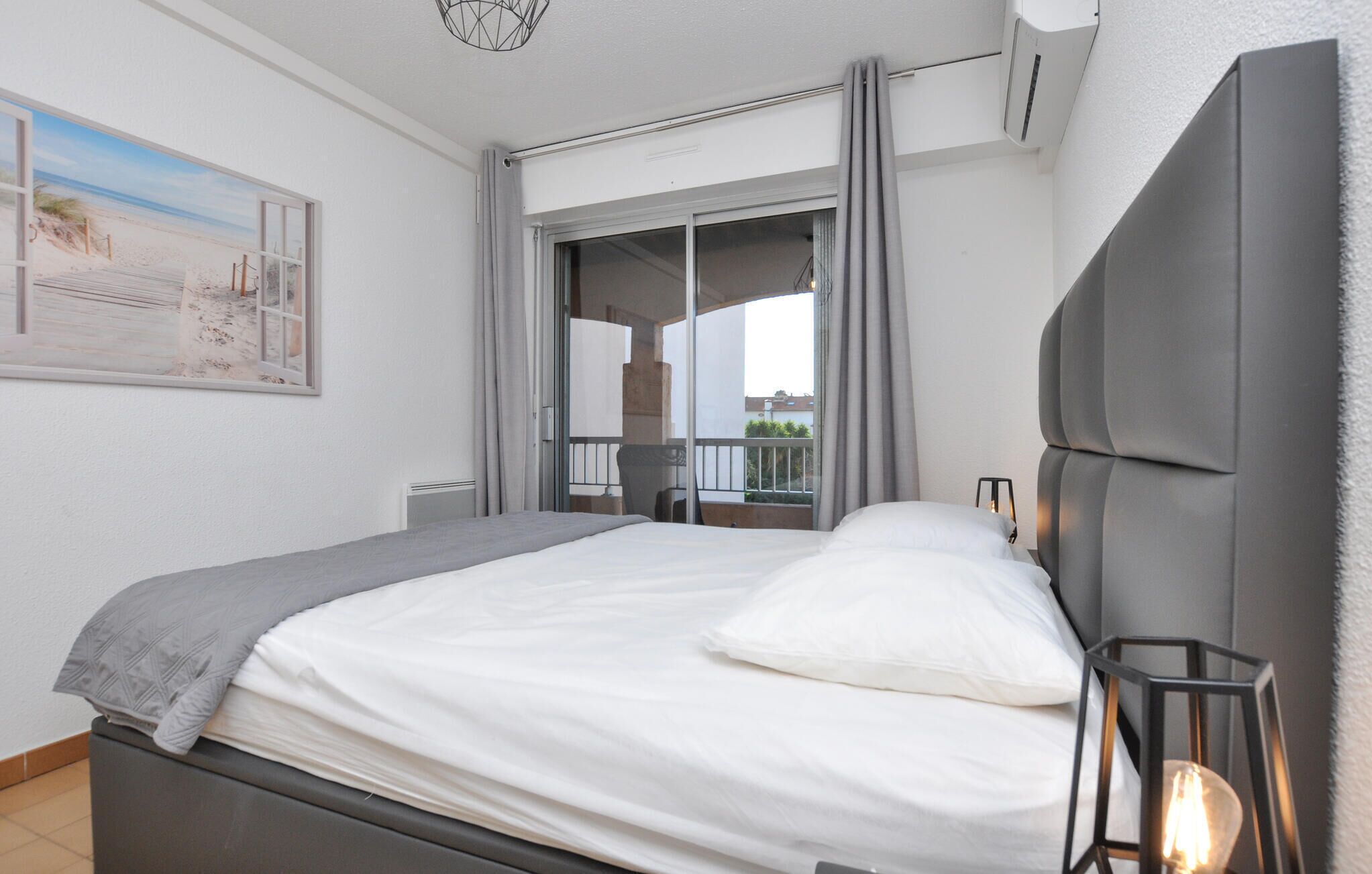 A bedroom with a bed, a nightstand, a lamp, and a view of the beach through a window.