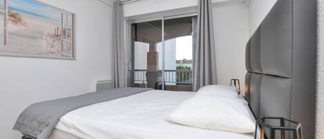 A bedroom with a bed, a nightstand, a lamp, and a view of the beach through a window.