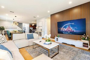 Una sala de estar moderna con un televisor de pantalla plana grande, un sofá blanco, una mesa de centro y un comedor al fondo.