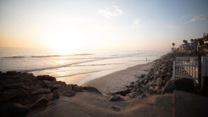 Una playa con costa rocosa, una valla blanca y edificios al fondo.