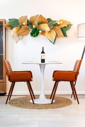 A dining area with a round table, two chairs, a bottle of wine, and a decorative wall piece.