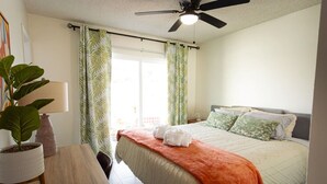 Un dormitorio con cama, ventilador de techo, ventana con cortinas y una planta en maceta.