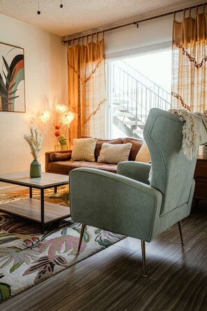 A modern living room with a light green armchair, a coffee table, and a sofa with cushions. There is a window with curtains, a staircase in the background, and a painting on the wall. - King Suite - Patio & Grill - Walk to Beach + Shops (Oceanside)