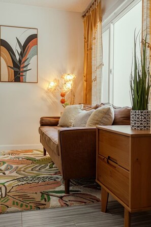 A cozy room with a sofa, a wooden cabinet, a patterned rug, and a wall-mounted light fixture. - King Suite - Patio & Grill - Walk to Beach + Shops (Oceanside)