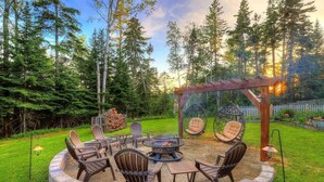 A circular fire pit area with seating, surrounded by a stone wall, set in a lush garden with trees and a wooden pergola. - Beautiful Spacious Well-Stocked Home in Downtown Rangeley. Pet & Family Friendly (Rangeley)