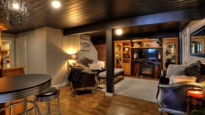 A cozy living room with a wooden floor, a bar area with stools, a fireplace, and a television. - Beautiful Spacious Well-Stocked Home in Downtown Rangeley. Pet & Family Friendly (Rangeley)