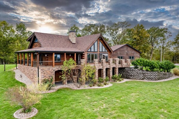 A large log cabin with a stone foundation, a covered porch, and a stone bridge.