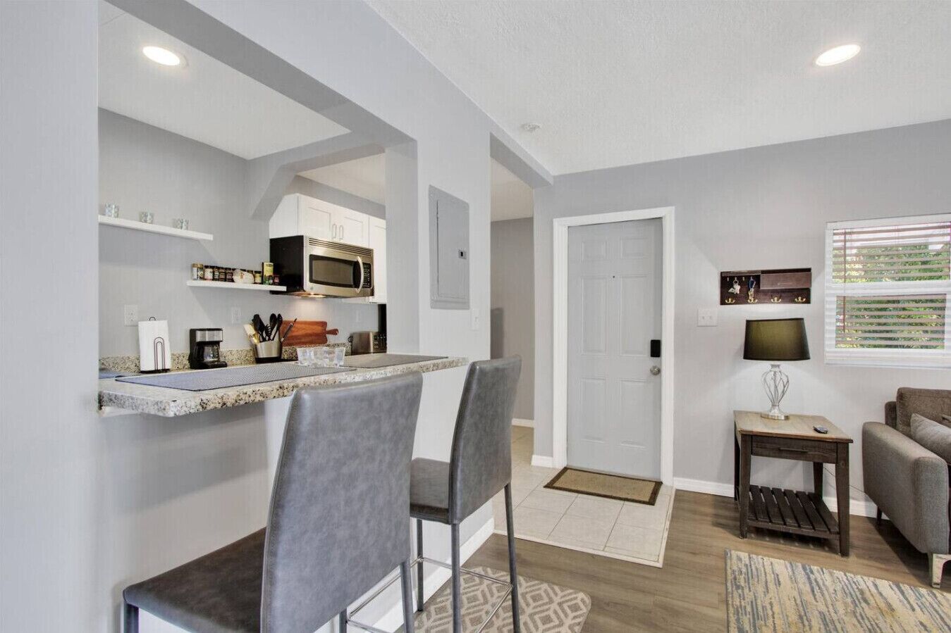 Un espacio habitable compacto con kitchenette, una zona de comedor con taburetes de bar, un pequeño rincón de estar con sofá y una ventana con persianas.