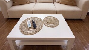 A beige sofa with three cushions, a white coffee table with two woven placemats and a remote control, and a sheer white curtain in the background.