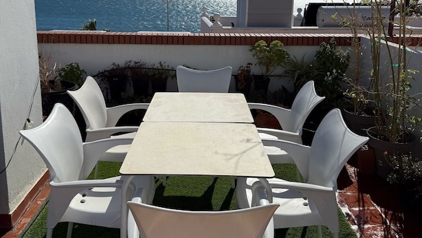 A patio with a table and chairs, overlooking a body of water, with a building and a fence in the background.
